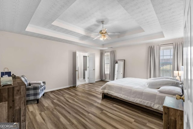 bedroom with a raised ceiling, a textured ceiling, wood finished floors, baseboards, and ceiling fan