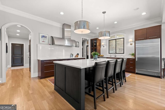 kitchen with arched walkways, light countertops, light wood-style floors, wall chimney range hood, and stainless steel built in refrigerator