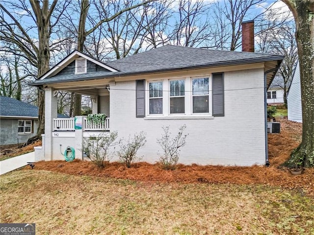 bungalow with a shingled roof, a chimney, cooling unit, a porch, and brick siding