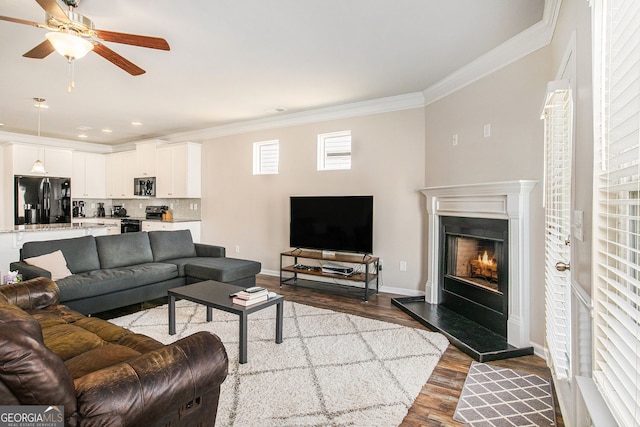 living room with baseboards, dark wood finished floors, ceiling fan, ornamental molding, and a lit fireplace