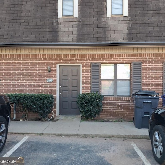 entrance to property with brick siding, uncovered parking, mansard roof, and roof with shingles