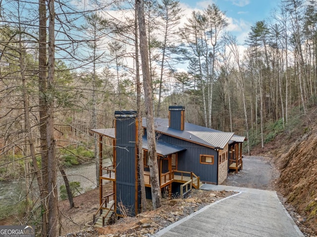 view of front of home featuring metal roof, driveway, a wooden deck, a chimney, and a wooded view