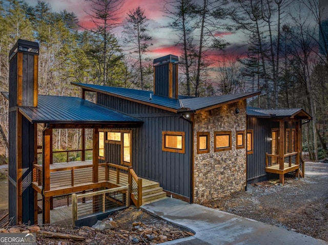 back of property at dusk featuring stone siding, a chimney, metal roof, and board and batten siding