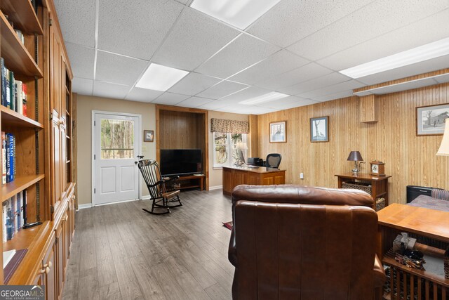 living area featuring wooden walls, baseboards, a drop ceiling, and wood finished floors