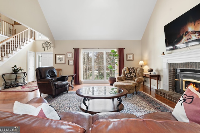 living area featuring high vaulted ceiling, wood finished floors, baseboards, stairs, and a brick fireplace