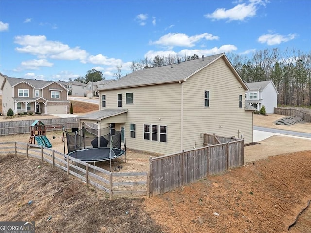 rear view of property featuring a trampoline, a playground, fence, and roof with shingles