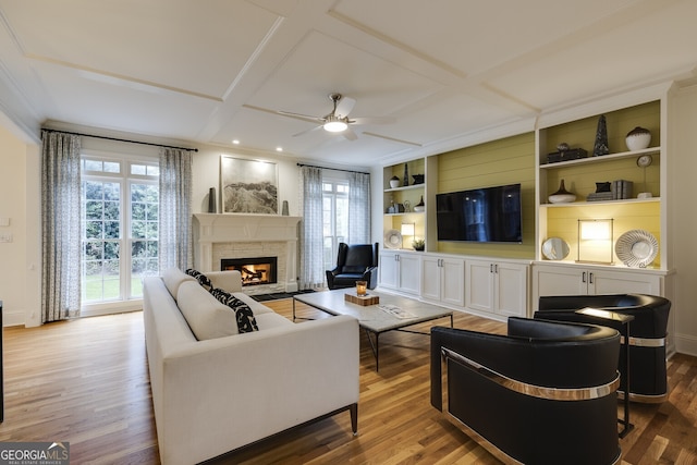 living area featuring built in features, a ceiling fan, wood finished floors, a warm lit fireplace, and coffered ceiling