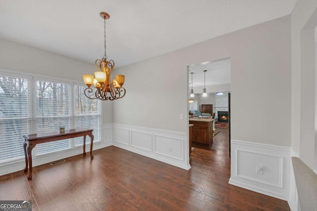 dining area featuring dark wood-type flooring, wainscoting, a lit fireplace, and an inviting chandelier