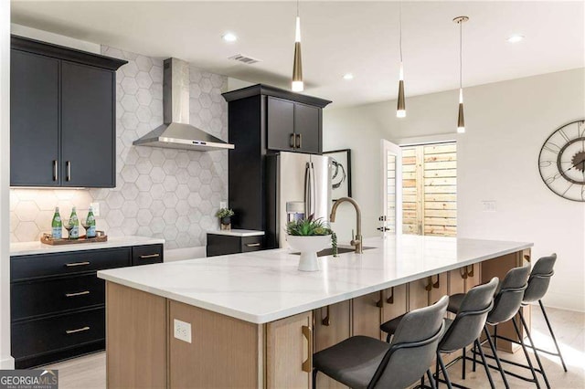 kitchen featuring stainless steel fridge with ice dispenser, backsplash, light stone countertops, wall chimney exhaust hood, and an island with sink