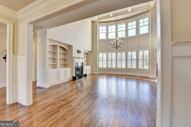 unfurnished living room with a glass covered fireplace, wood finished floors, crown molding, built in shelves, and a notable chandelier