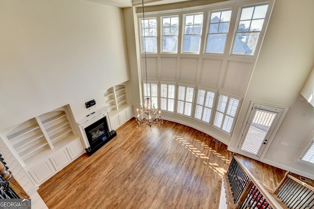 unfurnished living room featuring built in features, a towering ceiling, a glass covered fireplace, a chandelier, and light wood-type flooring