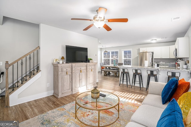 living area with visible vents, baseboards, light wood-style flooring, ceiling fan, and stairway