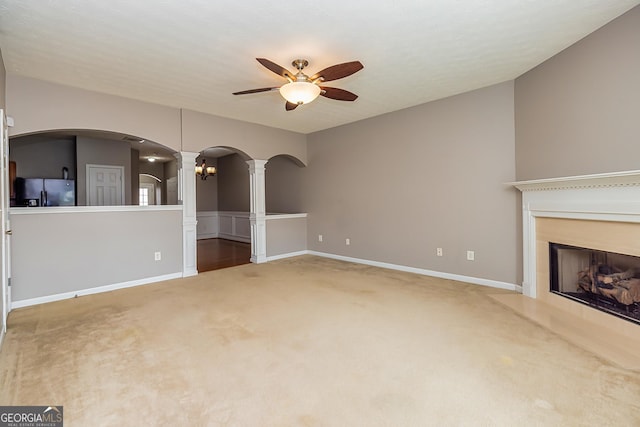 unfurnished living room featuring arched walkways, ceiling fan, a fireplace with raised hearth, carpet floors, and decorative columns