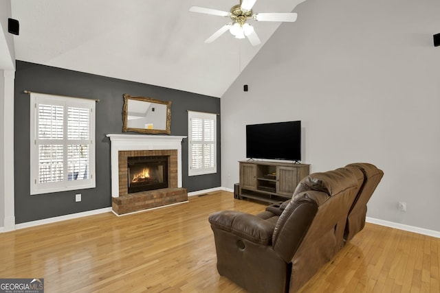 living area featuring baseboards, light wood-style flooring, ceiling fan, a fireplace, and high vaulted ceiling