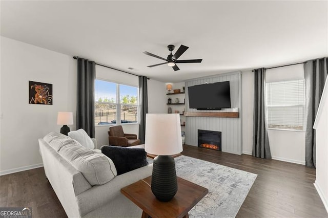 living area featuring a large fireplace, ceiling fan, baseboards, and dark wood finished floors