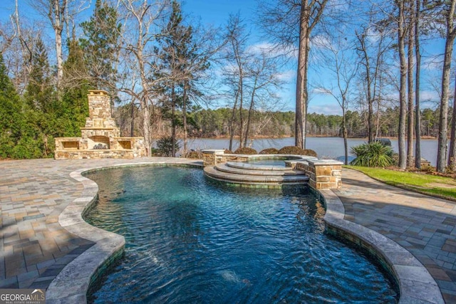 pool with an outdoor stone fireplace, a water view, a patio, and an in ground hot tub