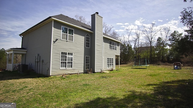 back of property with central air condition unit, a trampoline, a chimney, and a yard