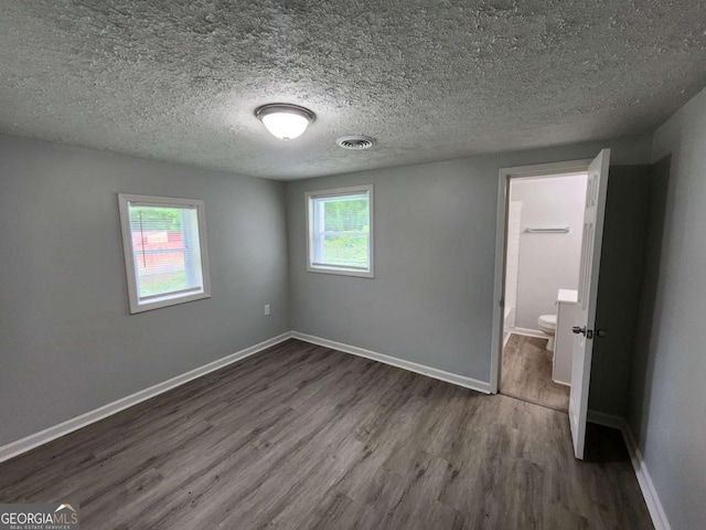 unfurnished bedroom with a textured ceiling, ensuite bathroom, dark wood-style flooring, visible vents, and baseboards