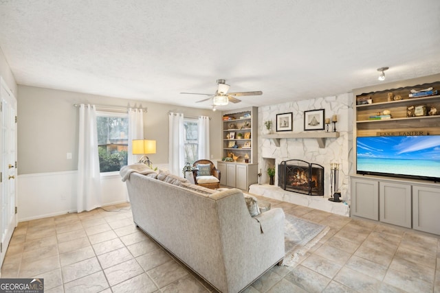 living room with a textured ceiling, ceiling fan, a stone fireplace, and built in shelves