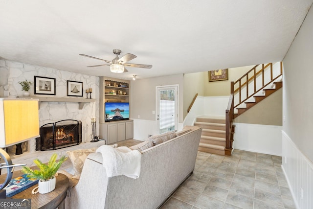 living room with built in features, a wainscoted wall, a fireplace, stairway, and ceiling fan