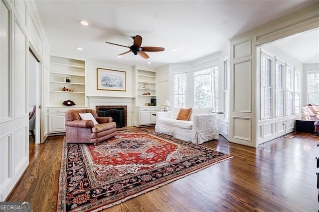 living area featuring dark wood-type flooring, built in shelves, a wealth of natural light, and ceiling fan