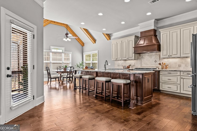 kitchen featuring premium range hood, dark wood-type flooring, backsplash, and an island with sink