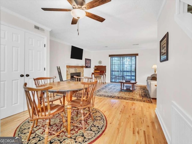 dining room featuring light wood-style flooring, a ceiling fan, visible vents, a tiled fireplace, and crown molding