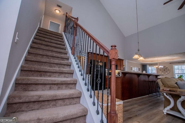 stairs with ceiling fan with notable chandelier, high vaulted ceiling, wood finished floors, and visible vents