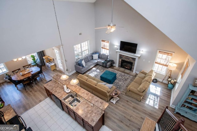 living room with ceiling fan, a stone fireplace, a high ceiling, baseboards, and light wood-style floors