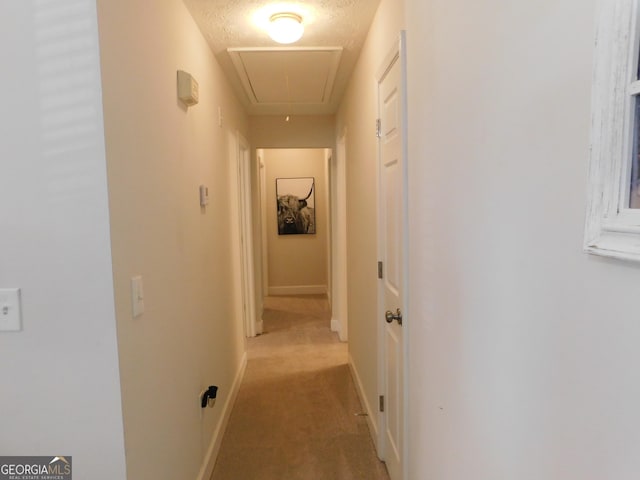 corridor with light carpet, a textured ceiling, attic access, and baseboards