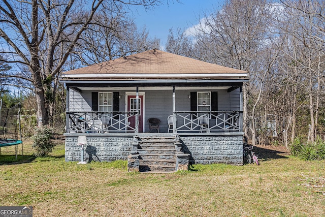 bungalow with a trampoline, covered porch, roof with shingles, and a front lawn