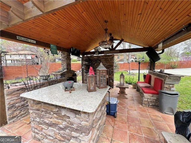 view of patio featuring a fenced backyard, an outdoor stone fireplace, a ceiling fan, outdoor dry bar, and a gazebo