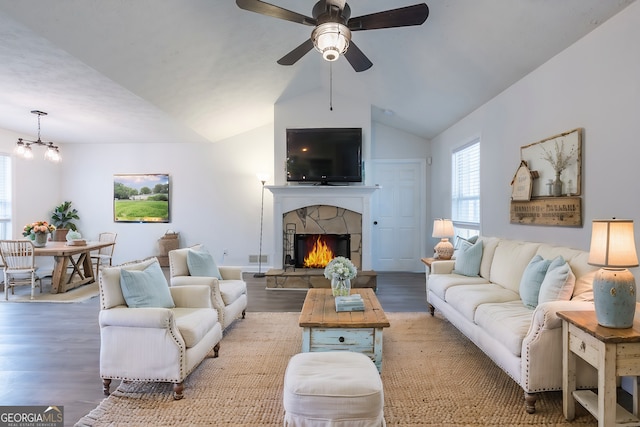 living room with ceiling fan with notable chandelier, vaulted ceiling, a stone fireplace, and wood finished floors