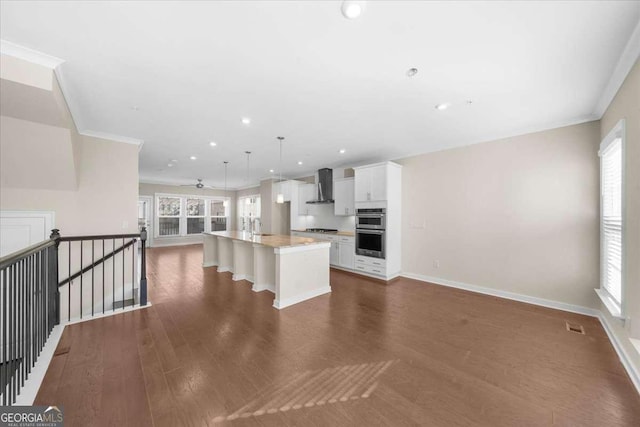 kitchen with ornamental molding, open floor plan, dark wood-type flooring, a kitchen island with sink, and wall chimney range hood