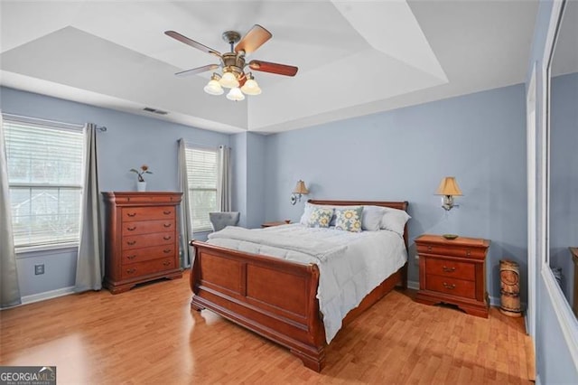 bedroom with light wood finished floors, baseboards, visible vents, a raised ceiling, and a ceiling fan