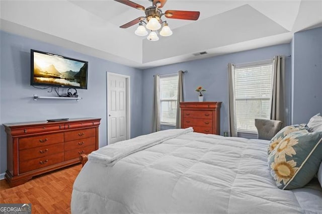 bedroom with a tray ceiling, light wood-style flooring, visible vents, and a ceiling fan