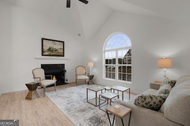 living area with ceiling fan, high vaulted ceiling, wood finished floors, a fireplace with flush hearth, and baseboards