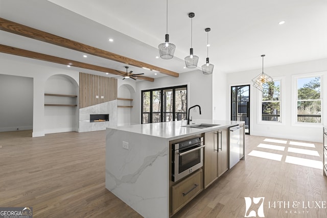 kitchen featuring stainless steel appliances, a high end fireplace, a sink, an island with sink, and light wood-type flooring