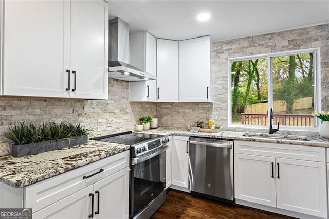 kitchen featuring appliances with stainless steel finishes, white cabinetry, a sink, light stone countertops, and wall chimney exhaust hood