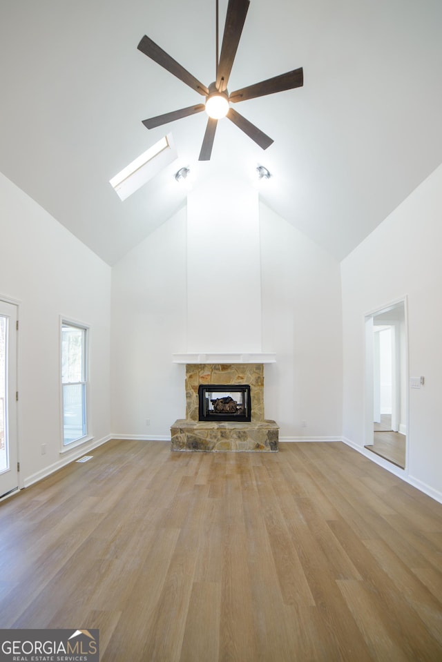 unfurnished living room with light wood finished floors, a stone fireplace, a skylight, high vaulted ceiling, and a ceiling fan