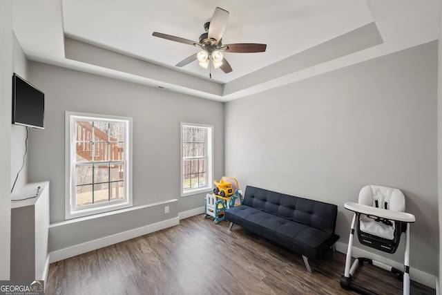 living area featuring a ceiling fan, a raised ceiling, baseboards, and wood finished floors