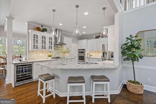 kitchen featuring beverage cooler, stainless steel appliances, a sink, white cabinetry, and wall chimney range hood