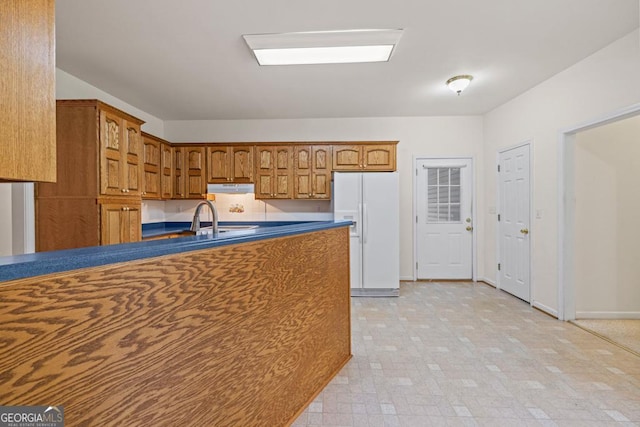 kitchen with dark countertops, white refrigerator with ice dispenser, brown cabinets, a sink, and under cabinet range hood