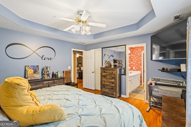 bedroom featuring a tray ceiling, visible vents, light wood-style flooring, ensuite bathroom, and a ceiling fan