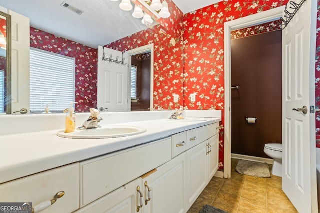 bathroom featuring toilet, double vanity, visible vents, and wallpapered walls