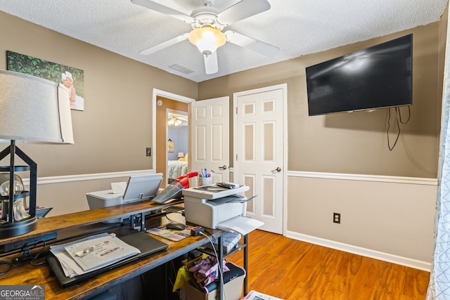 home office with a textured ceiling, wood finished floors, visible vents, baseboards, and a ceiling fan