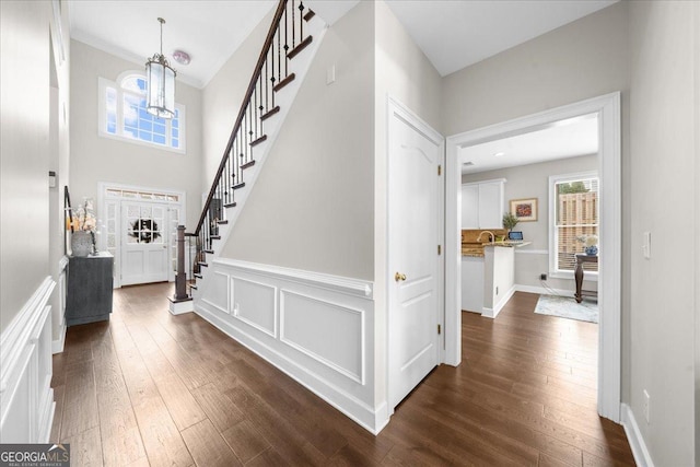 foyer with a wainscoted wall, stairway, dark wood-type flooring, a decorative wall, and a notable chandelier