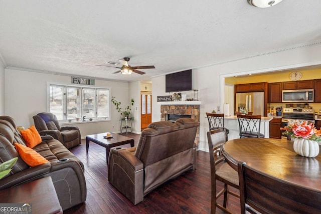 living room featuring dark wood finished floors, ceiling fan, ornamental molding, a textured ceiling, and a stone fireplace