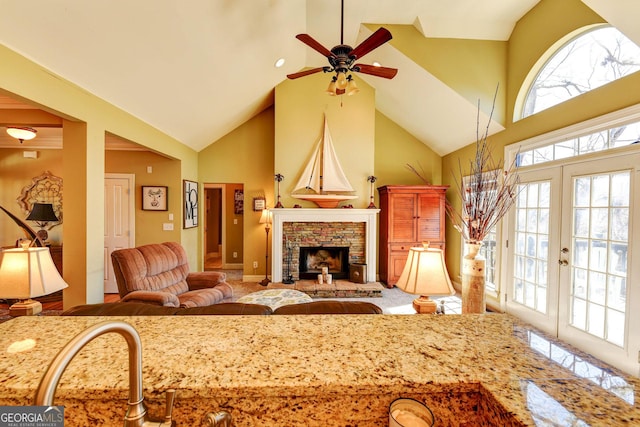 living area featuring a ceiling fan, high vaulted ceiling, french doors, and a stone fireplace