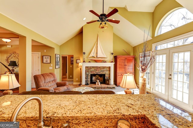 living area with high vaulted ceiling, plenty of natural light, a ceiling fan, a stone fireplace, and french doors